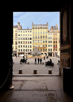 View of a public square as seen through an open door, that’s in shadow. There’s a decorative fountain, people milling about on foot and by motorbike.