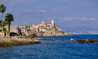 Antibes old town with pastel houses and stone towers above seaside ramparts, palm trees on the left.