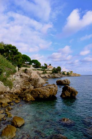 Boulders in clear turquoise water along a rocky coast, with pines and a villa on the headland.